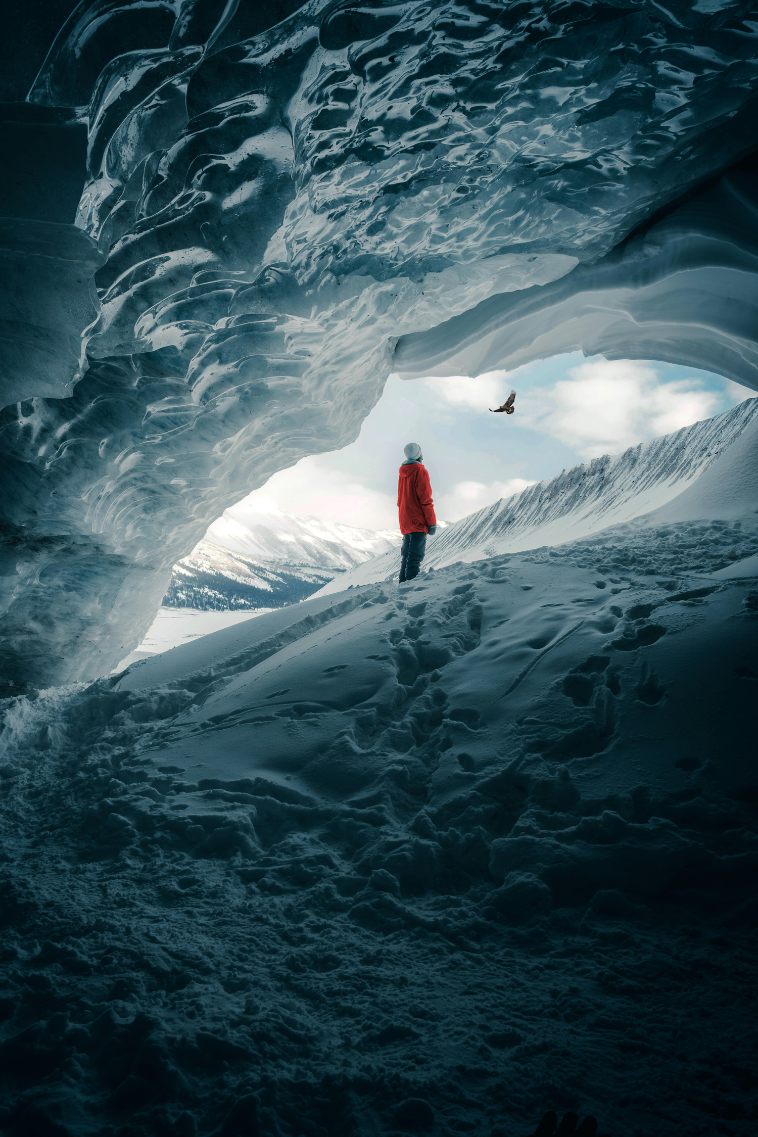 Person in a red jacket standing inside an ice cave with snow-covered mountains outside.