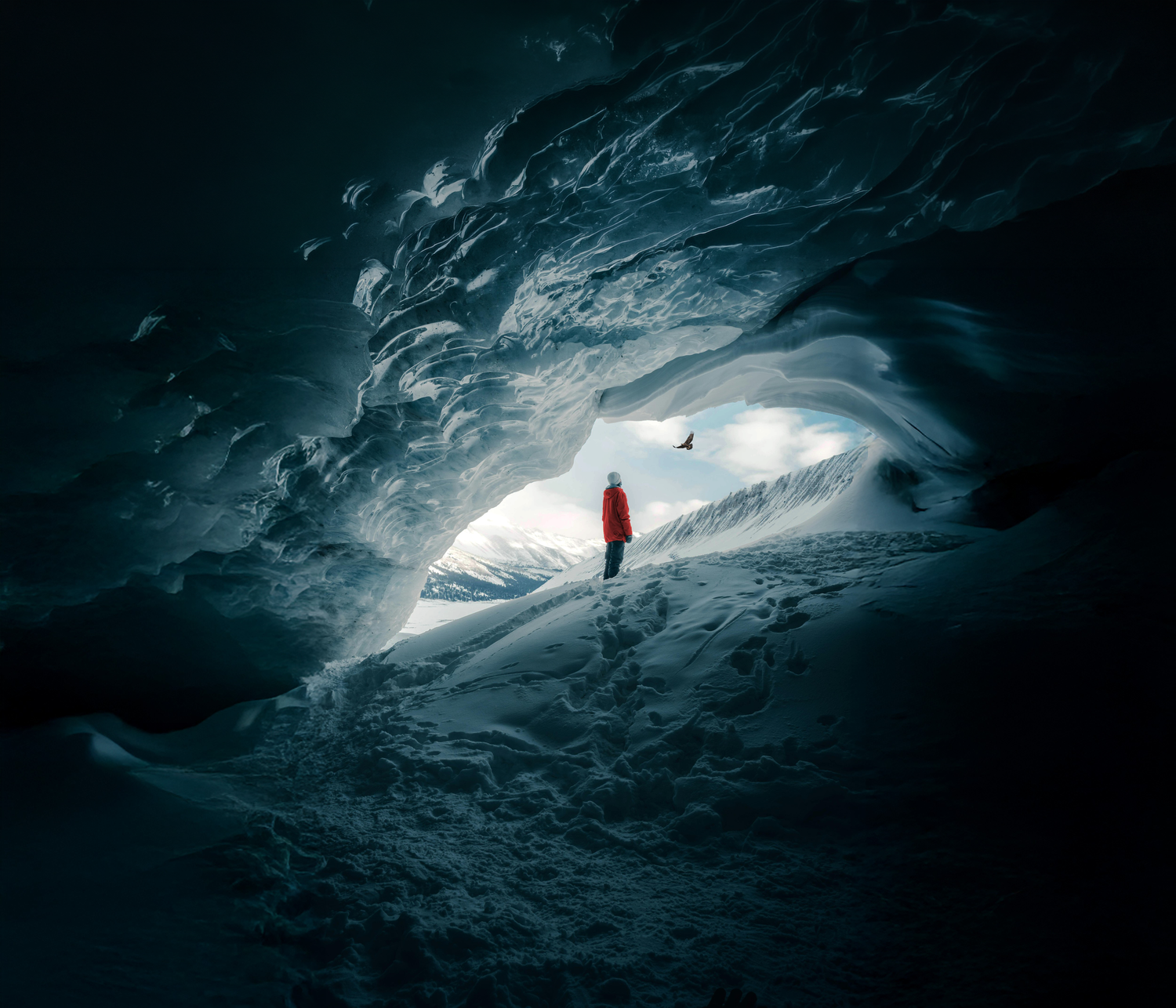 Person in a red jacket standing inside an ice cave with snow-covered mountains outside.