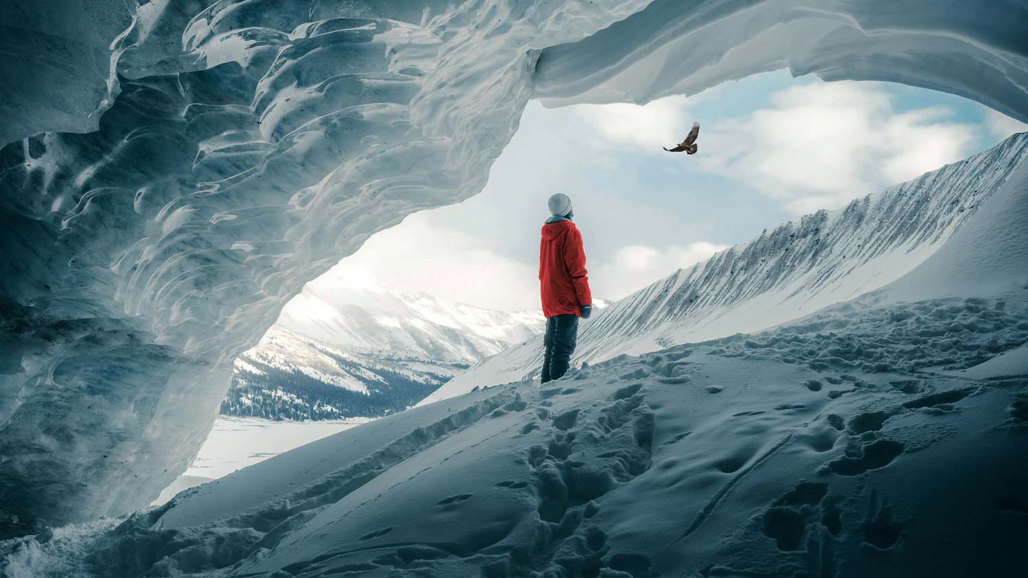Person in a red jacket standing inside an ice cave with snow-covered mountains outside.